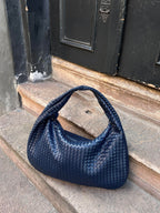 Sac à main tressé bleu marine posé sur des marches en pierre devant une porte noire. Accessoire mode élégant, tendance et chic.
