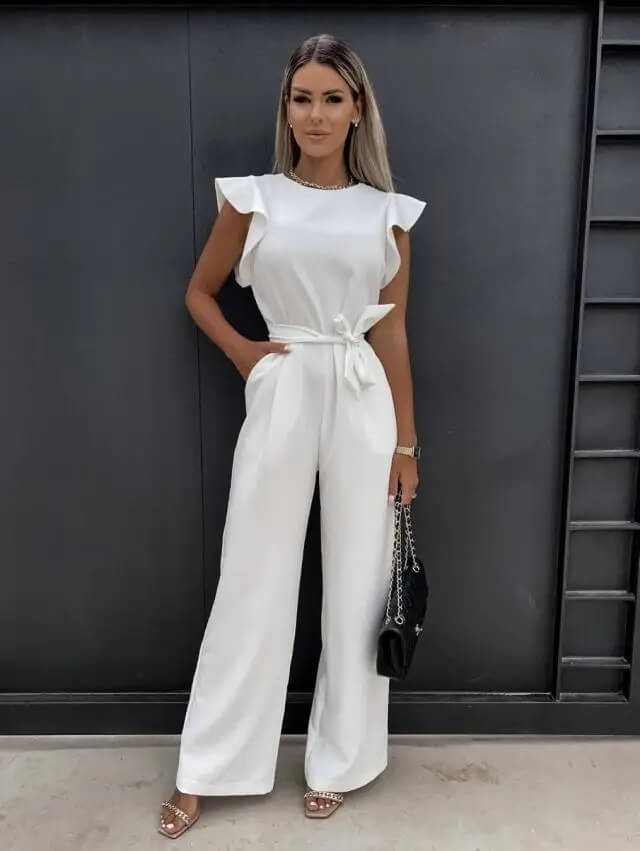 Femme portant une combinaison blanche élégante avec ceinture, posant devant un mur noir. Mode féminine chic, tenue de soirée tendance.