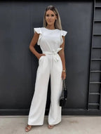 Femme portant une combinaison blanche élégante avec ceinture, posant devant un mur noir. Mode féminine chic, tenue de soirée tendance.