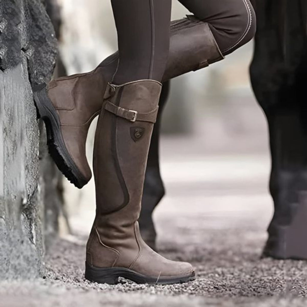 Bottes d'équitation marron en cuir, style équestre, portées par une personne en pantalon d'équitation, posées contre un mur en pierre.