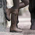 Bottes d'équitation marron en cuir, style équestre, portées par une personne en pantalon d'équitation, posées contre un mur en pierre.