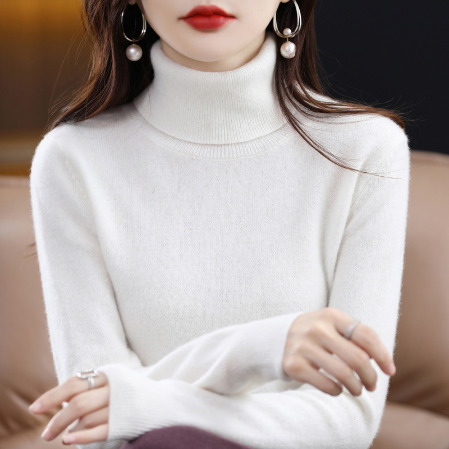 Femme portant un pull blanc à col roulé, boucles d'oreilles pendantes avec perles, assise sur un canapé beige. Mode élégante, style automne-hiver.