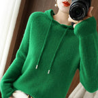 Femme portant un pull vert en laine avec capuche, tenant un appareil photo. Mode automne-hiver, style décontracté, photographie lifestyle.