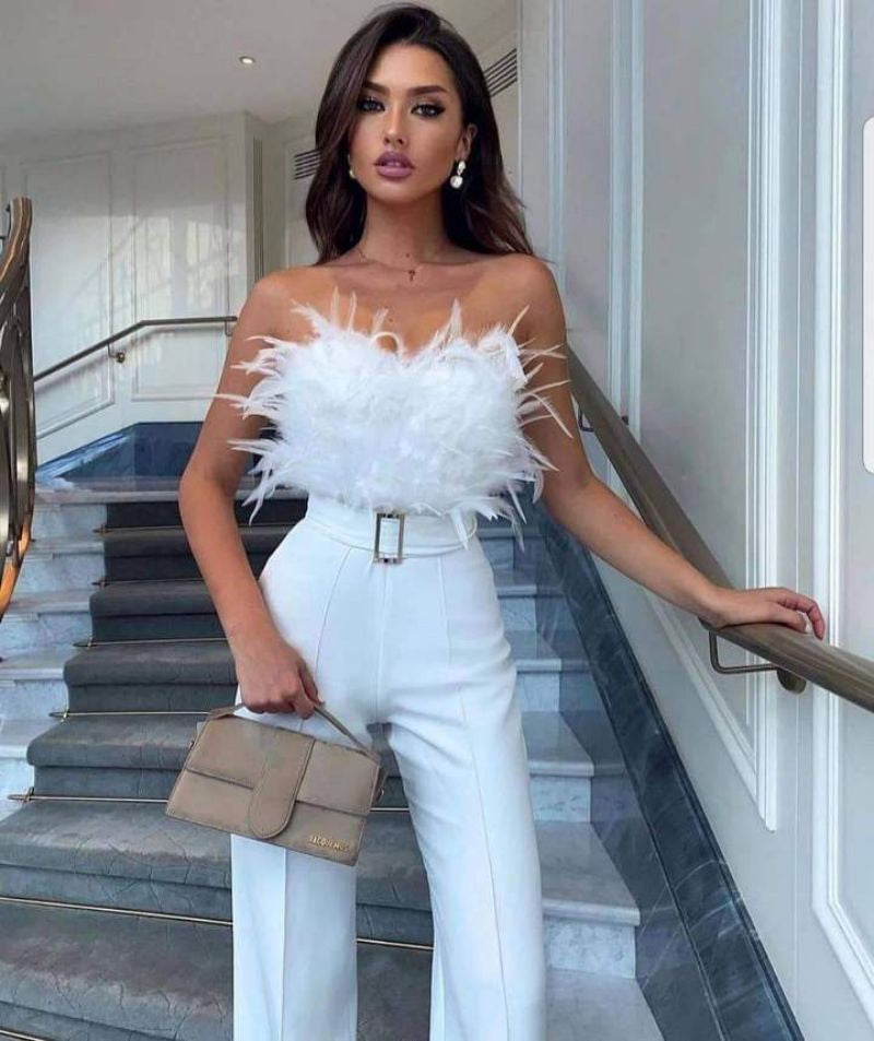 Femme élégante en tenue blanche chic, haut à plumes, pantalon taille haute, tenant un sac à main beige, posant dans un intérieur moderne.