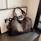 Sac à main en cuir marron foncé avec poches zippées, posé sur une table noire devant un cadre blanc. Accessoire mode élégant et pratique.
