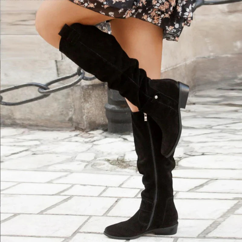 Bottes noires en daim pour femme, style cuissardes, portées avec une jupe fleurie. Mode automne, tendance chaussures élégantes.