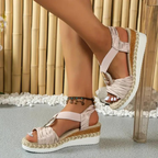 Sandales compensées rose doré pour femme, avec semelle en corde tressée et talon blanc, portées avec un bracelet de cheville, sur fond de bambou.