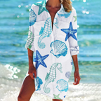 Robe de plage blanche avec motifs marins bleus et verts, incluant étoiles de mer, hippocampes et coquillages, portée par une femme au bord de la mer.