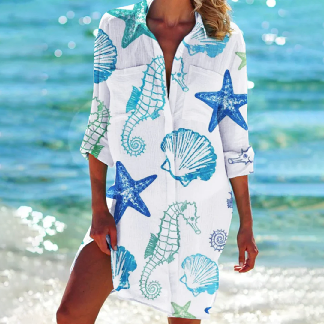 Robe de plage blanche avec motifs marins bleus et verts, incluant étoiles de mer, hippocampes et coquillages, portée par une femme au bord de la mer.
