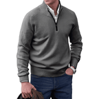 Homme portant un pull gris à col zippé, chemise blanche et jean bleu, tenant une veste marron. Mode masculine élégante et décontractée.