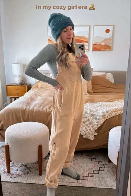 Femme en salopette beige et bonnet bleu prenant un selfie dans une chambre moderne, lit beige, décoration murale minimaliste. Mode décontractée, confort.