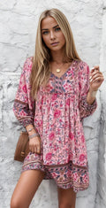 Femme en robe bohème rose à motifs floraux, posant devant un mur blanc texturé. Style chic et décontracté, tendance mode féminine.