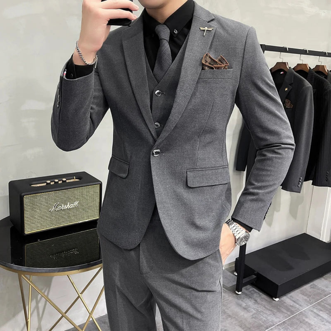 Homme en costume gris élégant, cravate noire, montre argentée, posant près d'une enceinte vintage et d'un portant à vêtements. Mode masculine chic.