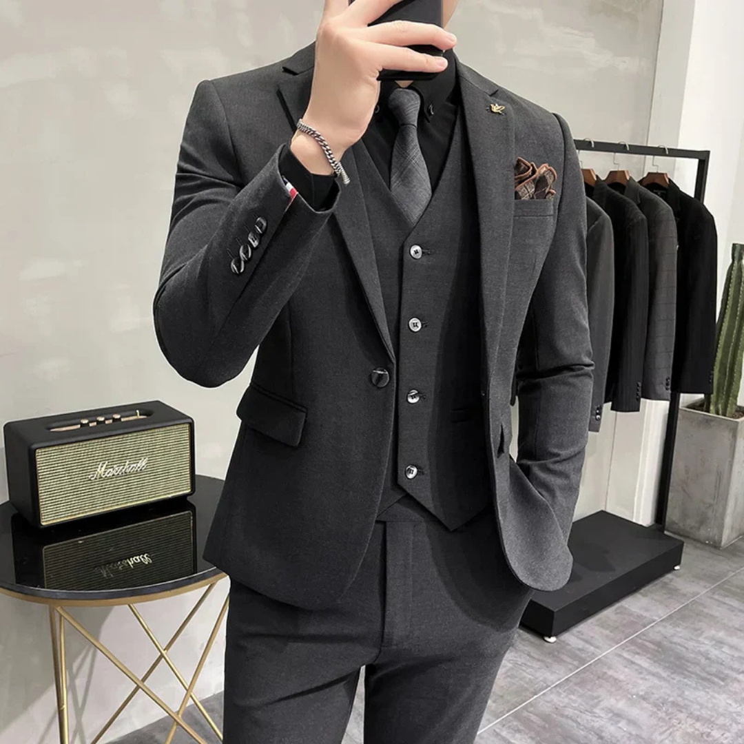 Homme en costume gris élégant, cravate noire, posant devant un amplificateur et un portant de vêtements. Mode masculine, style formel, boutique.