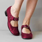 Chaussures rouges à talons bas avec boucle, portées par une personne debout. Design élégant et confortable, idéal pour mode féminine et occasions spéciales.