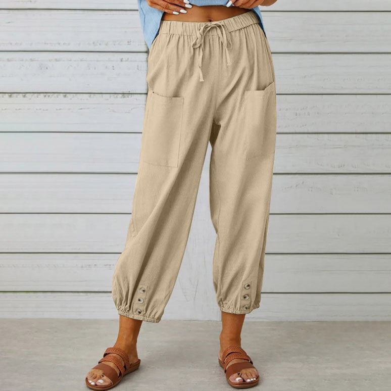 Pantalon beige ample pour femme avec poches avant, taille élastique et boutons aux chevilles, porté avec des sandales marron sur fond de mur en bois.