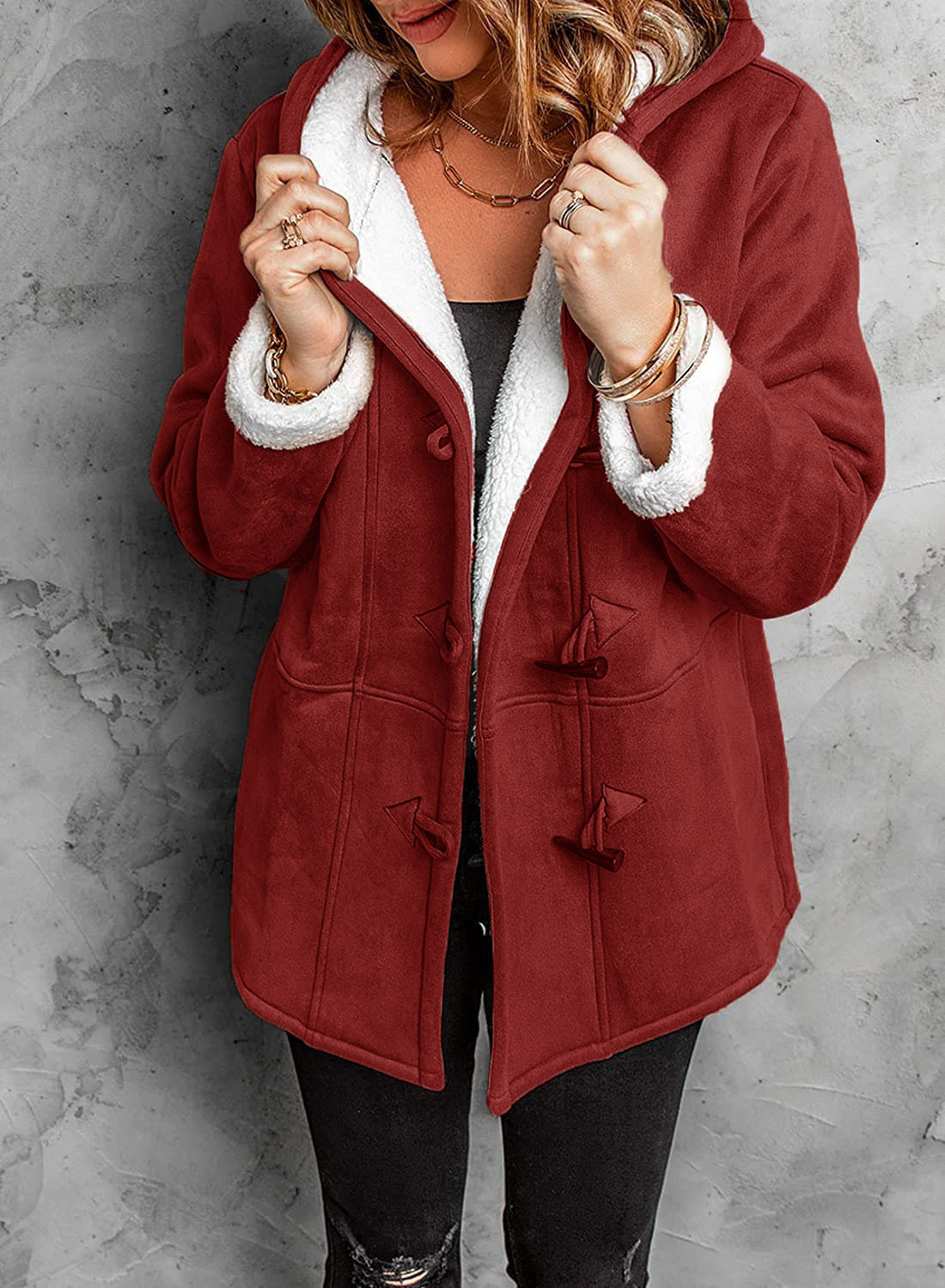 Femme portant un manteau rouge à capuche doublé de fourrure blanche, fermetures à brandebourgs, sur fond de mur gris. Mode hiver élégante et confortable.