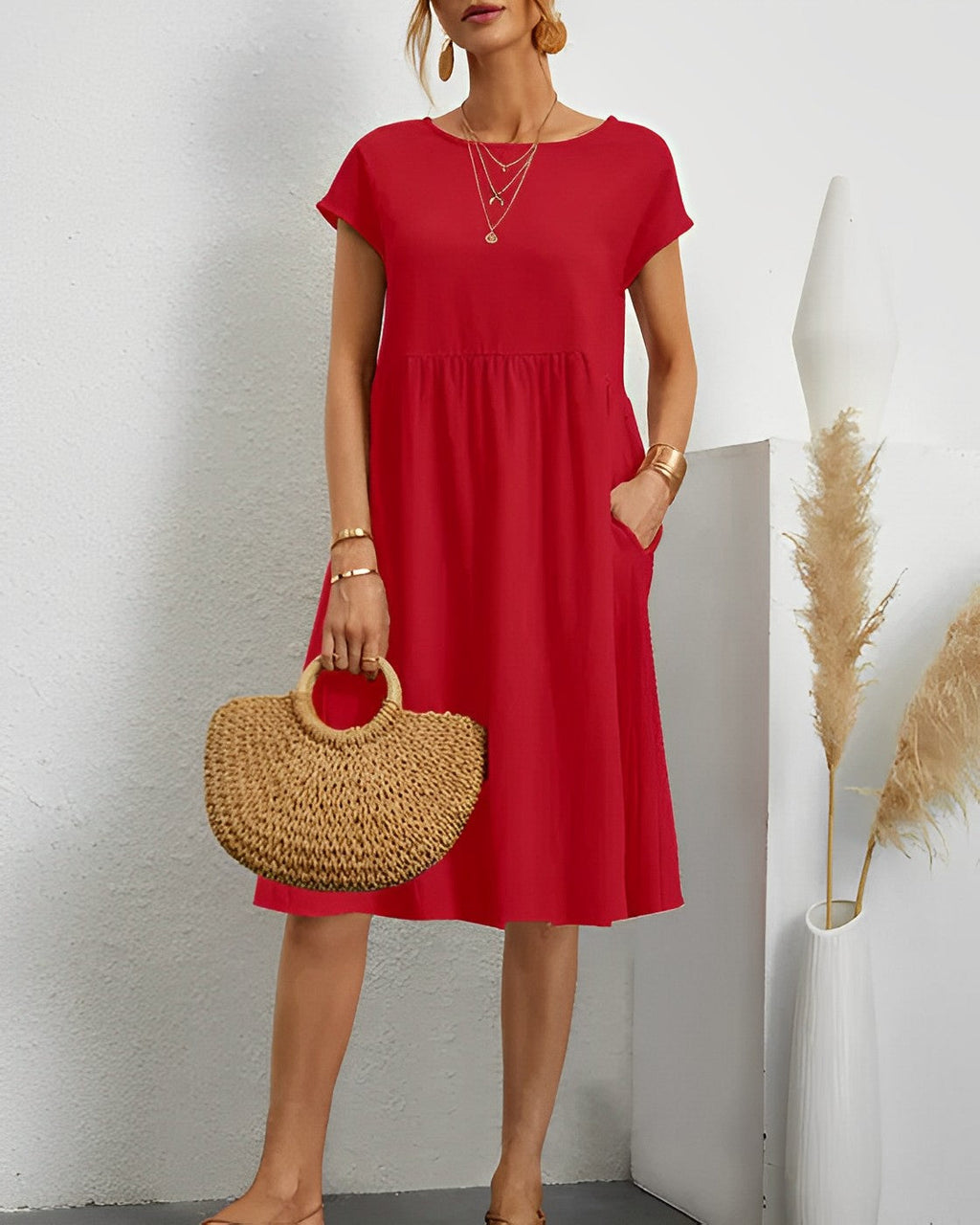 Robe rouge élégante avec manches courtes, portée par une femme tenant un sac en paille. Décor minimaliste avec vase blanc et herbes séchées.