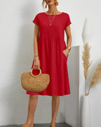 Robe rouge élégante avec manches courtes, portée par une femme tenant un sac en paille. Décor minimaliste avec vase blanc et herbes séchées.
