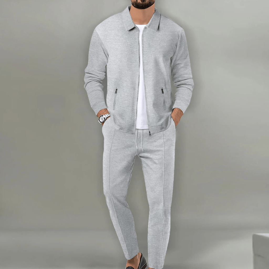 Homme portant un ensemble de survêtement gris élégant, composé d'une veste zippée et d'un pantalon assorti, sur fond neutre. Mode masculine moderne.