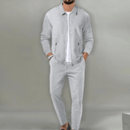 Homme portant un ensemble de survêtement gris élégant, composé d'une veste zippée et d'un pantalon assorti, sur fond neutre. Mode masculine moderne.