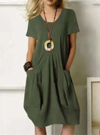Robe verte ample avec poches, portée par une femme. Accessoires: collier pendentif rond, bracelets en bois. Mode décontractée chic.
