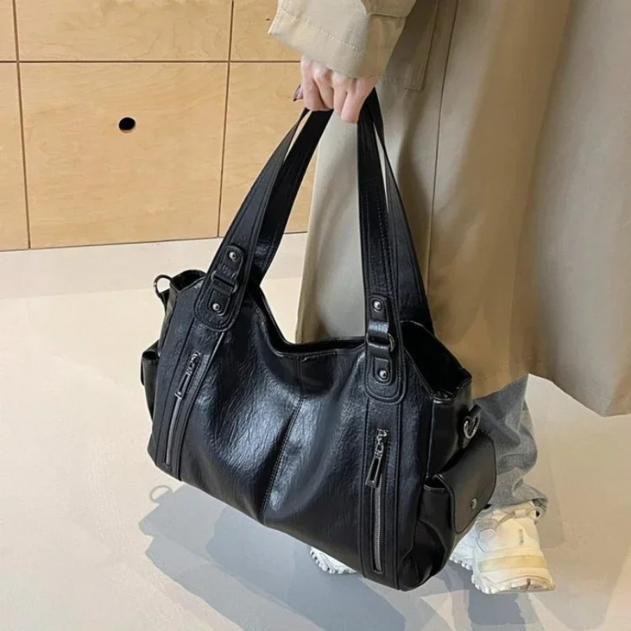 Sac à main noir en cuir porté par une personne en manteau beige, avec des poches zippées et des anses robustes, sur fond de sol clair et mur en bois.