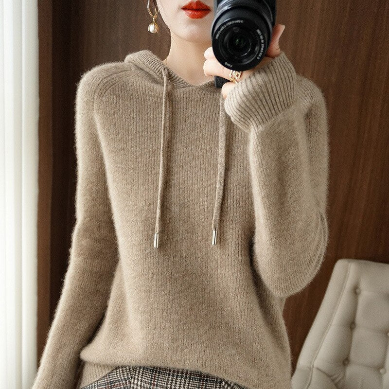 Femme en pull en cachemire beige, tenant un appareil photo, posant devant un fond en bois. Mode automne, style décontracté, élégance moderne.