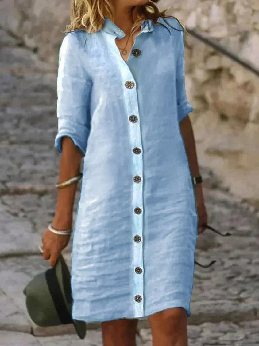 Robe chemise en lin bleu clair avec manches retroussées, boutons décoratifs, portée par une femme tenant un chapeau, sur fond de rue pavée.