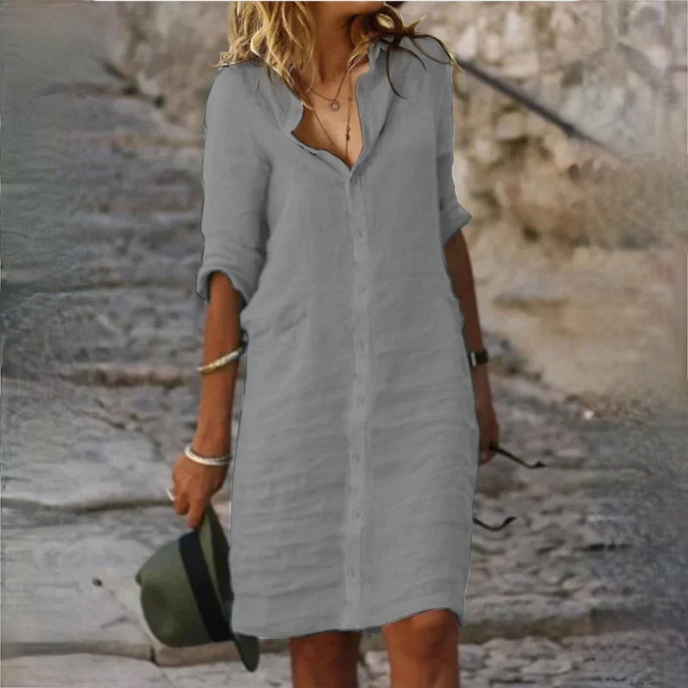 Robe chemise grise en lin pour femme, manches longues retroussées, tenue décontractée estivale, accessoirisée de bracelets et chapeau vert.