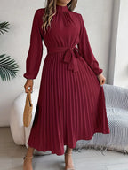 Robe longue plissée bordeaux avec ceinture, manches longues, tenue élégante pour femme, accessoire sac en paille, intérieur moderne.