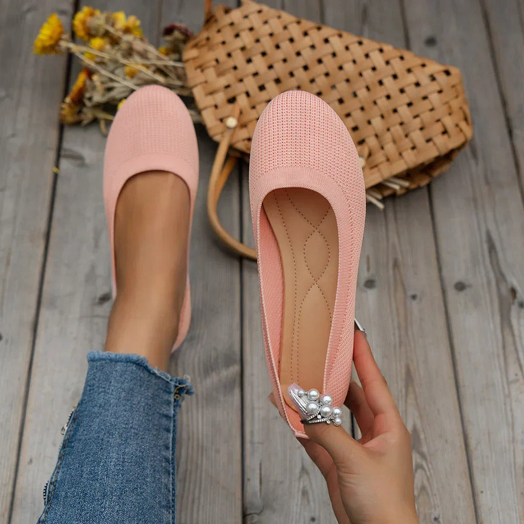 Ballerines roses en tissu texturé, tenues par une main, posées sur un sol en bois avec un sac en osier et des fleurs jaunes en arrière-plan.