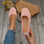 Ballerines roses en tissu texturé, tenues par une main, posées sur un sol en bois avec un sac en osier et des fleurs jaunes en arrière-plan.