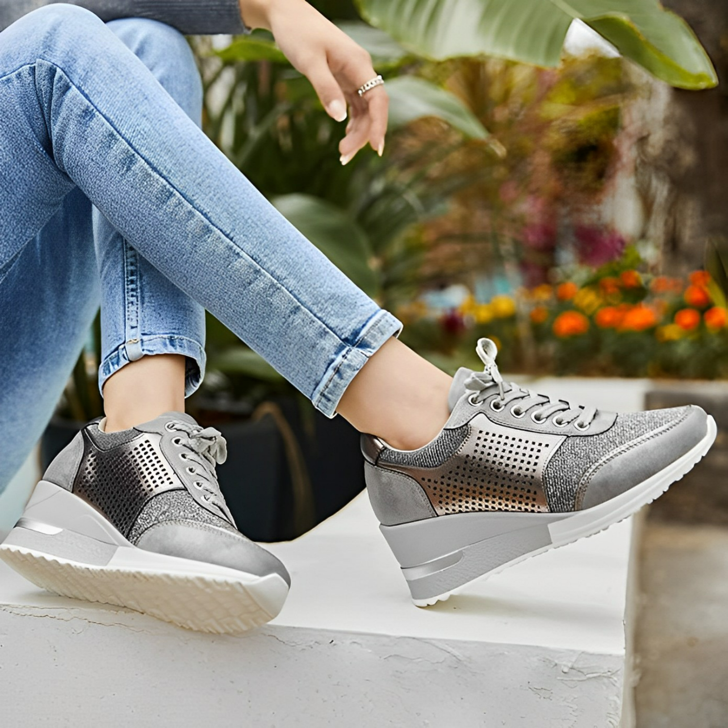 Baskets argentées pour femme avec semelle compensée, portées avec un jean bleu clair, posées sur un muret, fond de plantes et fleurs colorées.