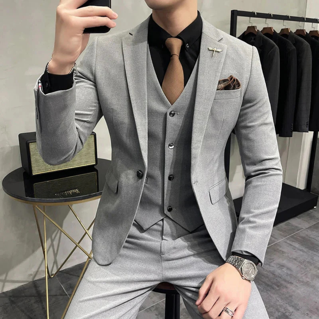 Homme en costume gris élégant, cravate marron, chemise noire, montre argentée, assis près d'une table moderne, vêtements suspendus en arrière-plan.