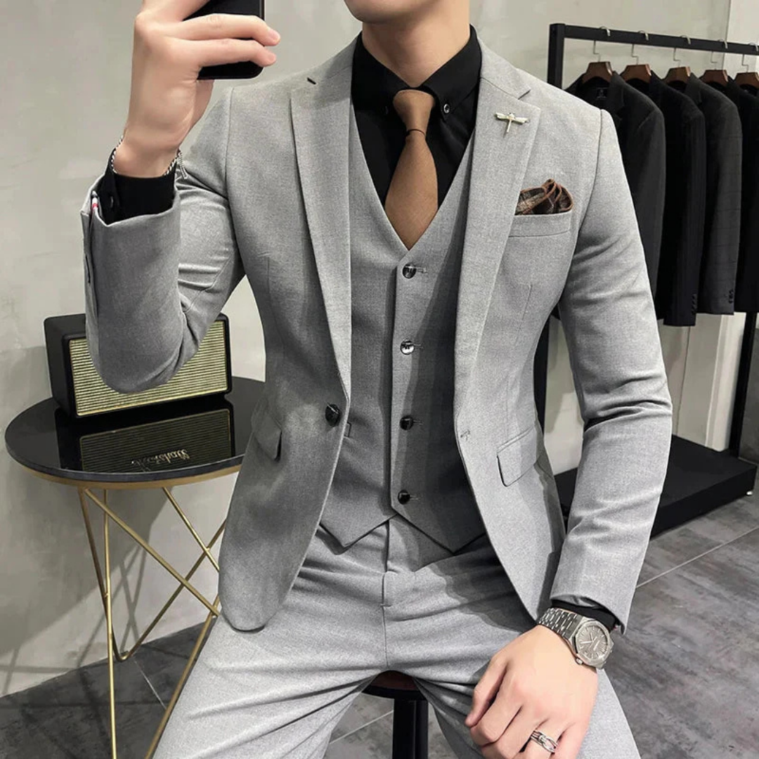 Homme en costume gris élégant, cravate marron, chemise noire, montre argentée, assis près d'une table moderne, vêtements suspendus en arrière-plan.