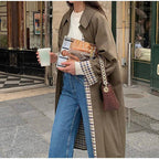 Femme en manteau marron tenant un café et un magazine, portant un sac à main marron et un jean, marchant dans une rue urbaine.