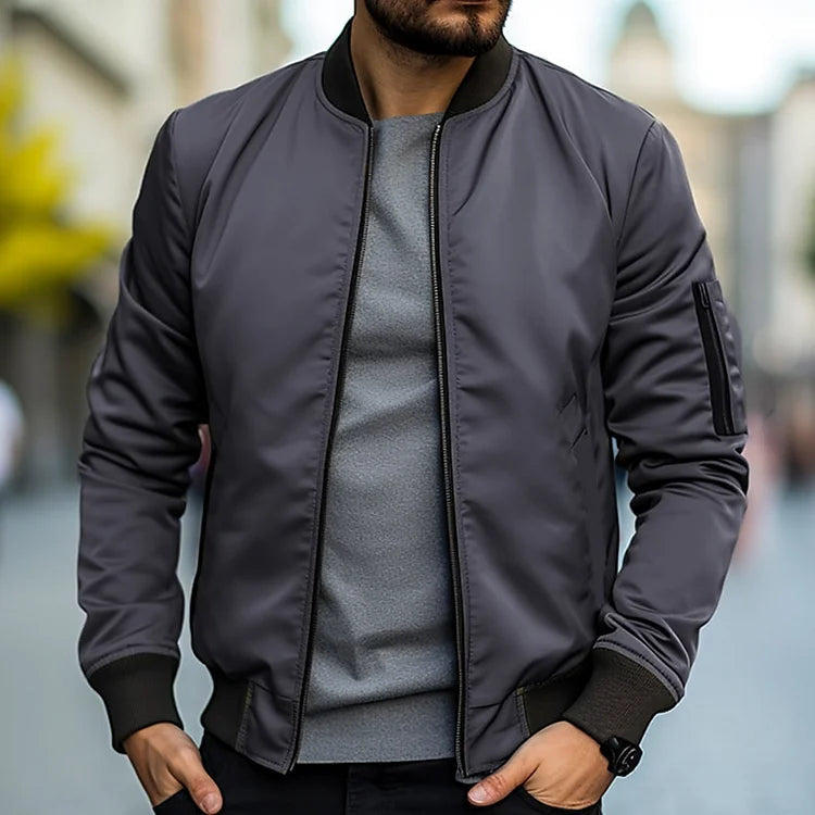 Homme portant un blouson bomber gris foncé, t-shirt gris, dans une rue urbaine floue. Mode masculine, style décontracté, tendance automne.