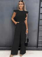 Femme portant une combinaison noire élégante avec volants, posant contre un mur gris. Mode féminine chic, tenue de soirée tendance.