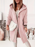 Femme portant un manteau rose en tricot avec capuche, sac blanc à bandoulière, posant devant des affiches. Mode automne-hiver, style décontracté.
