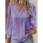 Femme portant un chemisier violet plissé avec manches longues et col en V, accessoirisé de colliers dorés, devant un mur en briques blanches.