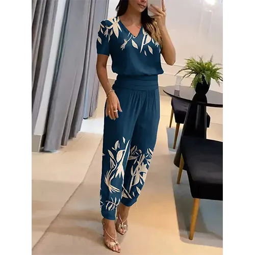 Femme portant une combinaison bleue à motifs floraux blancs, debout dans un salon moderne. Mode élégante, tenue décontractée chic, style estival.