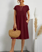 Robe bordeaux élégante avec manches courtes, accessoires dorés, sac en paille, décor minimaliste, pampas en vase blanc. Mode féminine chic.