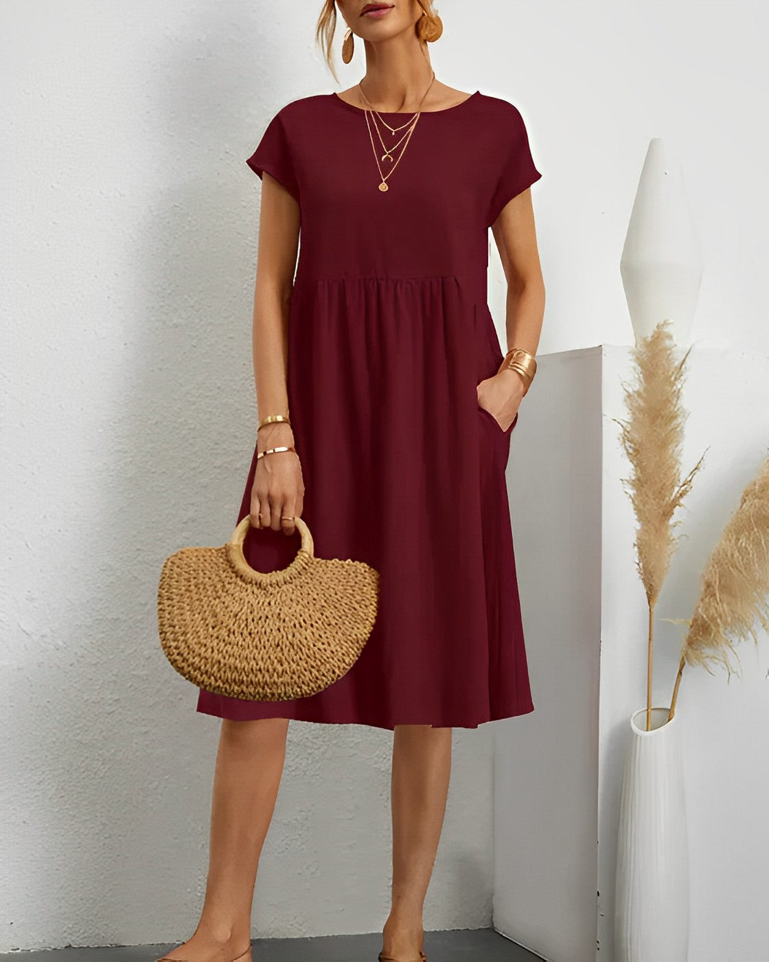 Robe bordeaux élégante avec manches courtes, accessoires dorés, sac en paille, décor minimaliste, pampas en vase blanc. Mode féminine chic.