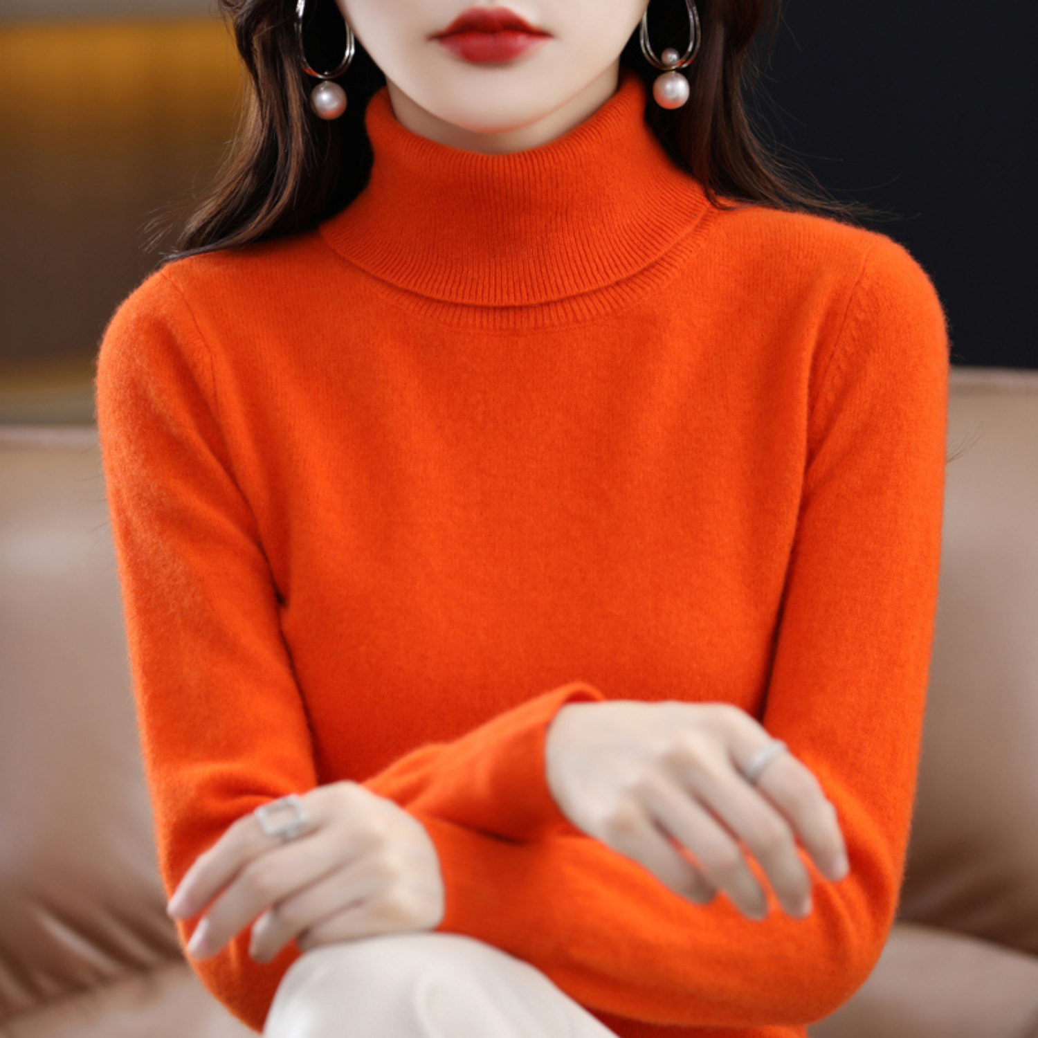 Femme portant un pull col roulé orange vif, assise sur un canapé en cuir. Accessoires: boucles d'oreilles perles, bague argentée. Mode automne-hiver.