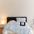 Lit moderne avec couverture texturée bleue, tablette affichant une image, lampe design sur table de chevet, ambiance minimaliste et cosy.