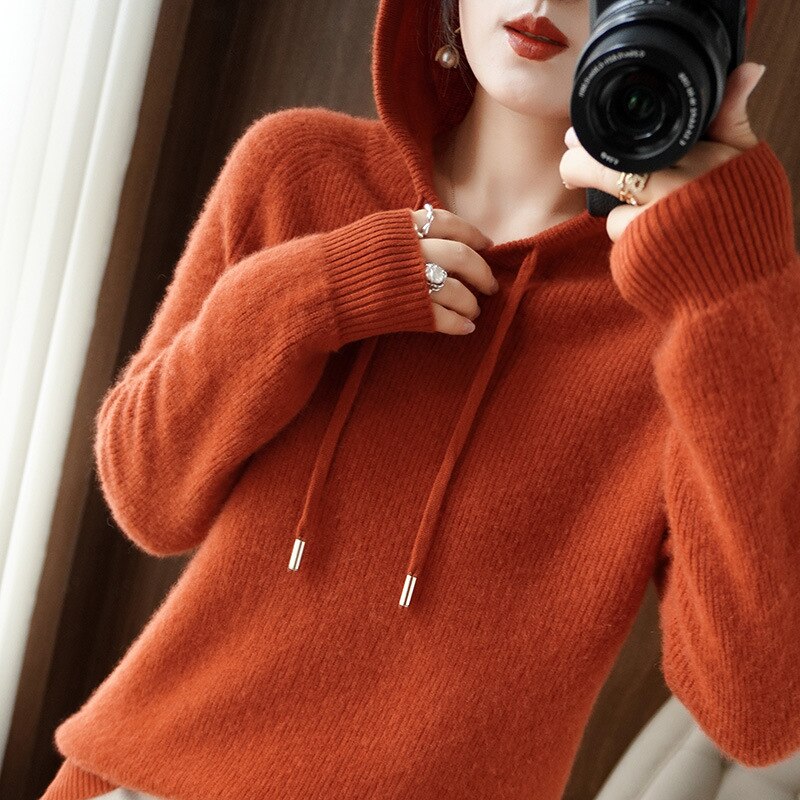 Femme en pull à capuche orange, tenant un appareil photo. Mode automne, style décontracté, photographie intérieure.
