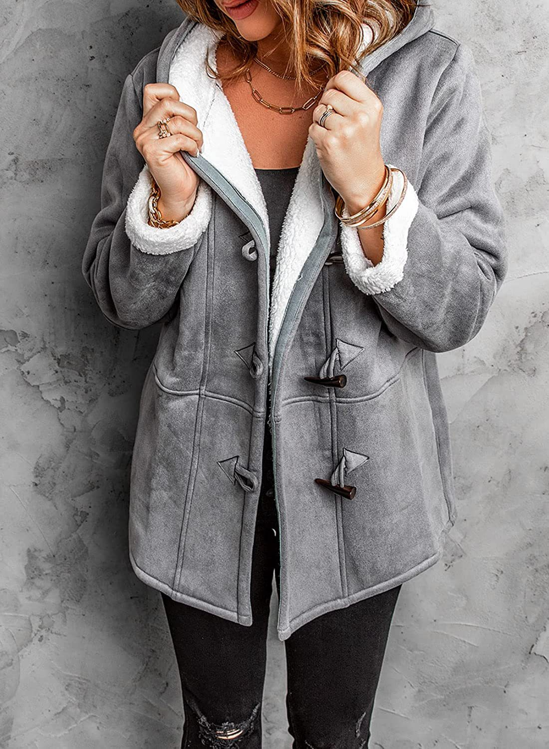 Femme portant un manteau en laine grise avec doublure en fourrure blanche, boutons en bois, sur fond de mur gris. Mode hiver élégante et confortable.