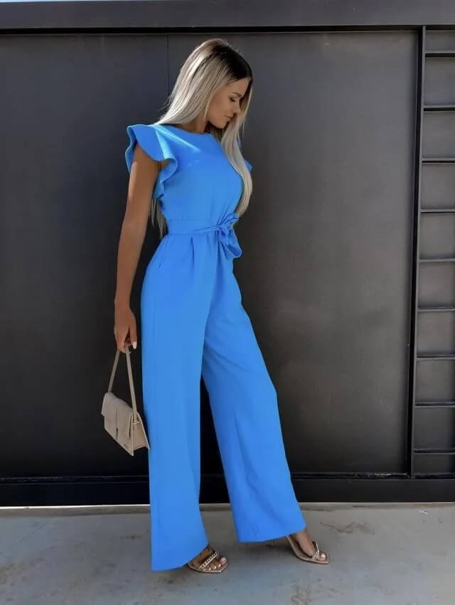 Femme en combinaison bleue élégante, tenant un sac à main beige, posant devant un mur noir. Mode féminine, style chic, tenue d'été.