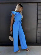 Femme en combinaison bleue élégante, tenant un sac à main beige, posant devant un mur noir. Mode féminine, style chic, tenue d'été.